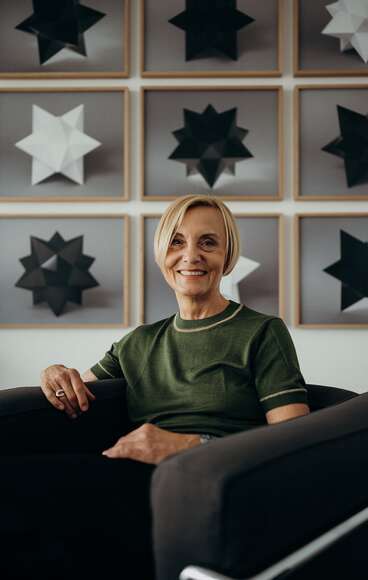 A person in a green shirt sits on a chair, smiling, with geometric star art on the wall behind them.