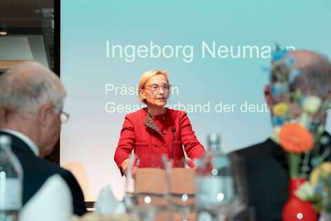 Eine Frau in rotem Blazer hält eine Rede bei einer Jahrestagung, umgeben von Zuhörern und Blumenarrangements.