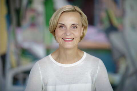 A smiling woman with short blonde hair wears a white top against a colorful, blurred background.