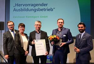 A group of five people stands on stage, smiling, as they present an award certificate and bouquet at a ceremony.
