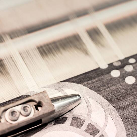 Ertex - BUSINESS AREAS - Fabric A close-up of a jacquard loom weaving a detailed geometric pattern on fabric with intricate white and gray designs.