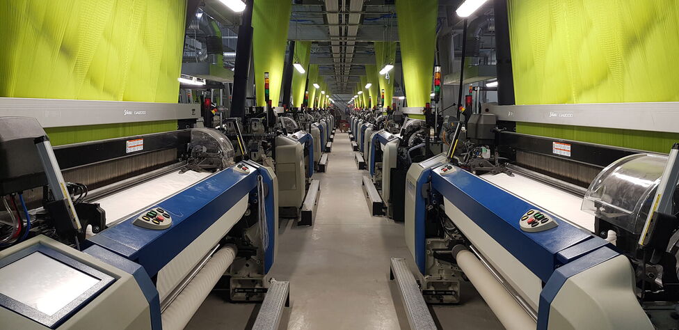 Rows of advanced textile machines in a factory are busy weaving fabric under bright lights.