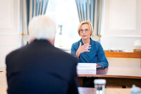 A woman in a blue jacket speaks to a man in a formal meeting setting, with a nameplate reading "Frau Neumann" visible.