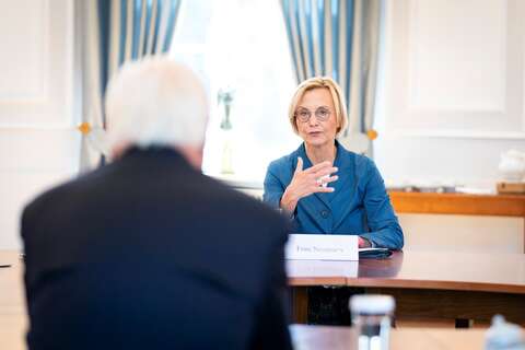 Eine Frau in einer blauen Jacke spricht gestikulierend mit einer anderen Person in einem Besprechungsraum.