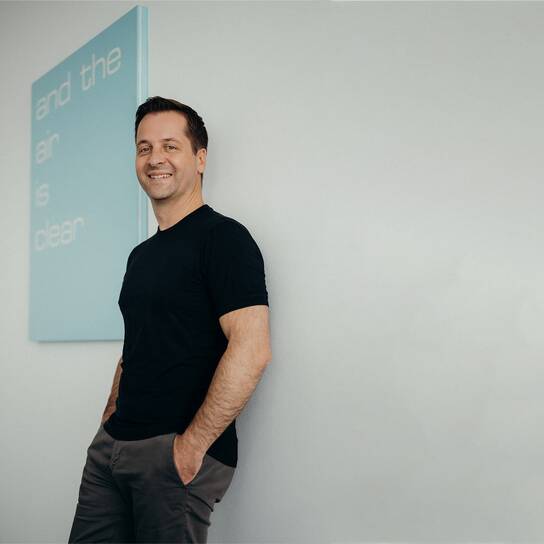 A man in a black shirt leans against a wall, smiling, with a blue sign reading "and the air is clear" in the background.