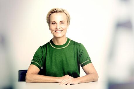 Eine Frau in einem grünen T-Shirt sitzt lächelnd an einem Tisch und strahlt historische Gelassenheit aus.
