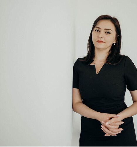 A woman in a black dress stands confidently against a white wall, conveying a sense of professionalism and poise.