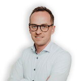 Ein Mann mit Brille und weißem Hemd posiert freundlich mit verschränkten Armen vor einem weißen Hintergrund.