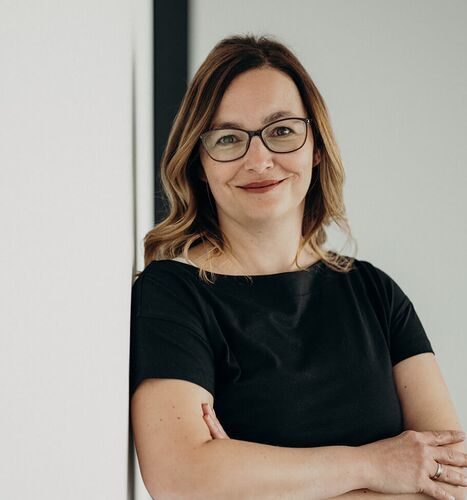 Eine lächelnde Frau mit Brille lehnt entspannt an einer weißen Wand und trägt ein schwarzes Kleid.