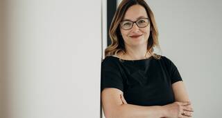 Eine lächelnde Frau mit Brille lehnt entspannt an einer weißen Wand und trägt ein schwarzes Kleid.