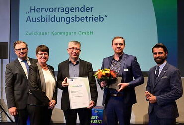 Eine Gruppe von fünf Personen posiert auf einer Bühne, während eine Person eine Auszeichnung und Blumen hält.