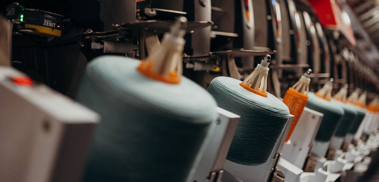 Rows of spinning machines with blue yarn spools are operating in a textile factory.