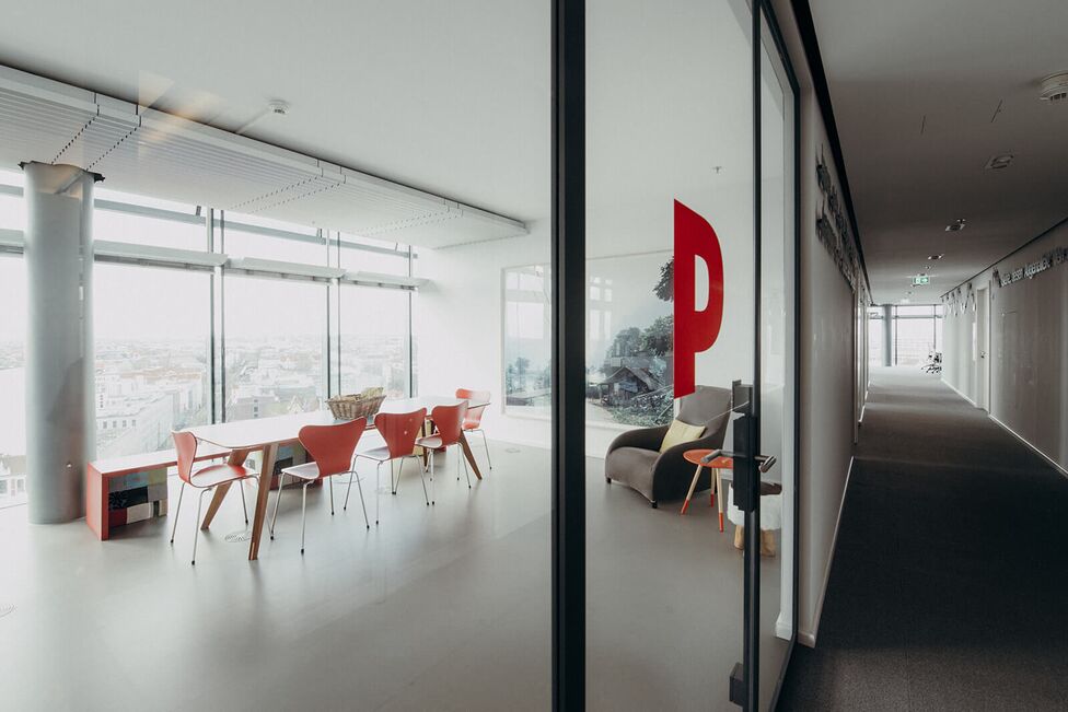 Ein moderner Konferenzraum mit großen Fenstern und roten Stühlen neben einem langen, lichtdurchfluteten Flur.