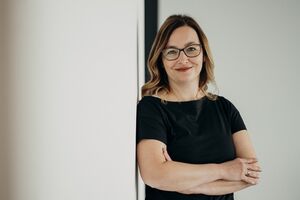 A woman in glasses and a black dress stands confidently with arms crossed, leaning against a white wall.