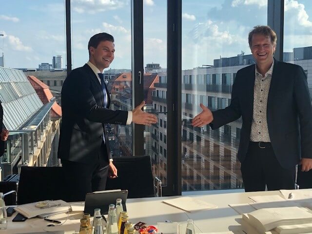 Two men in suits stand smiling, about to shake hands in a bright office with a city view.