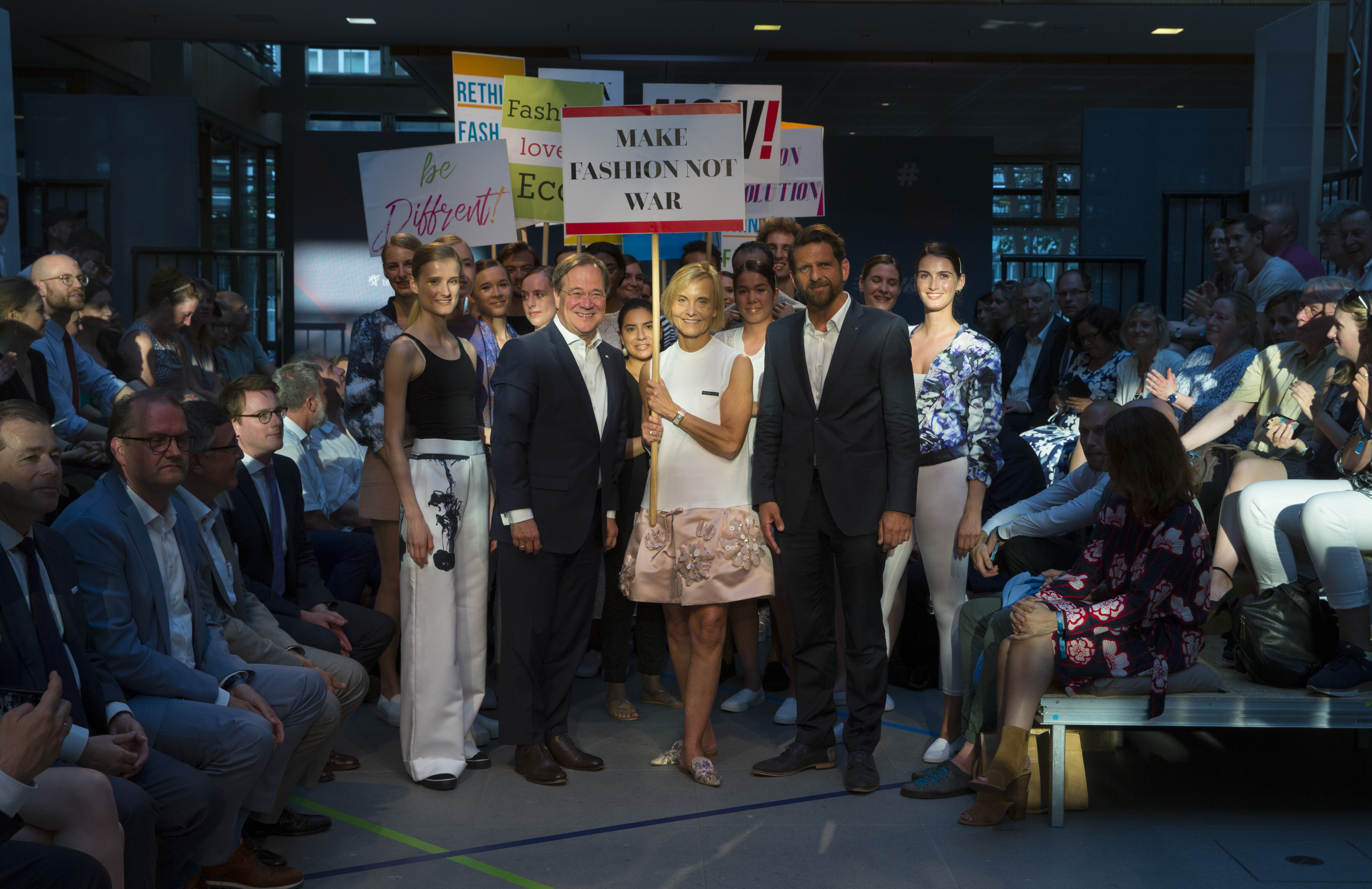Eine Gruppe von Menschen posiert mit Schildern wie "Make Fashion Not War" auf einem Mode-Event.